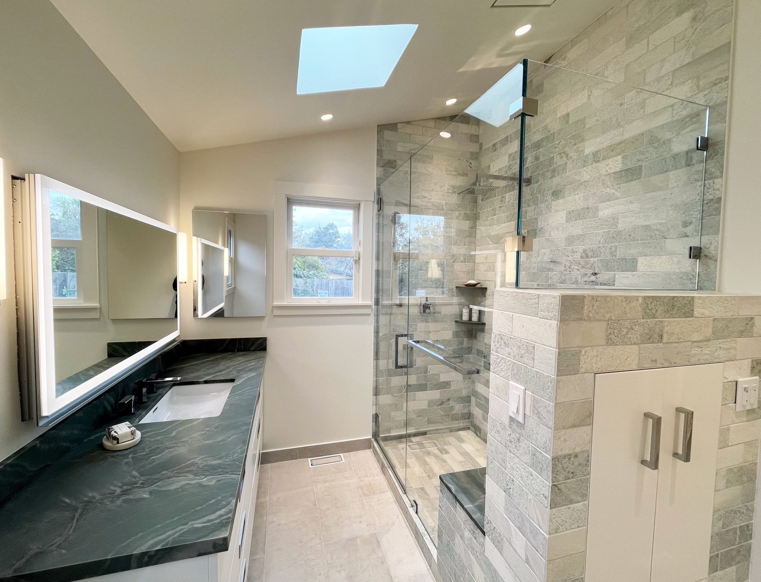 Remodeled bathroom with stone subway tile shower, dark stone vanity, and skylight